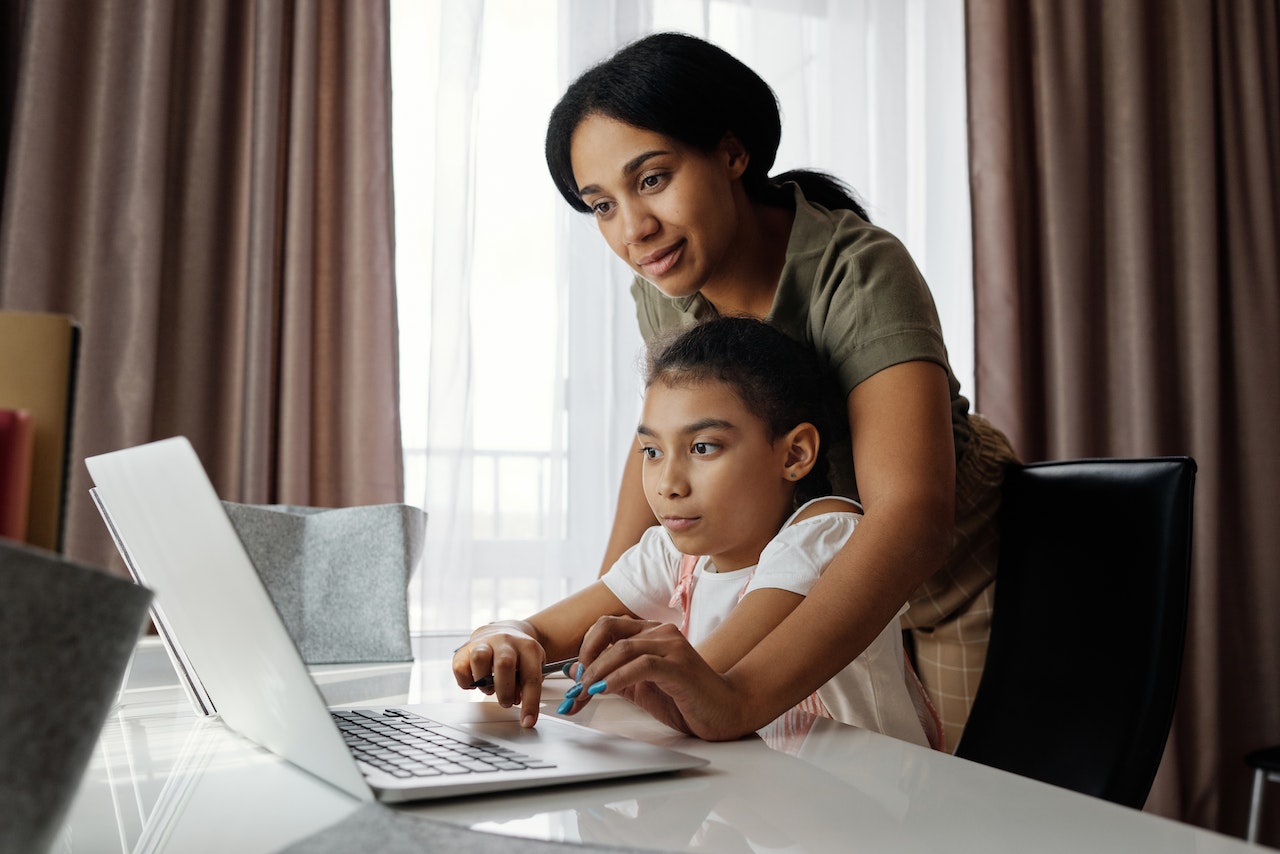 Mother standing behind her child sitting in front of a laptop on a white table.